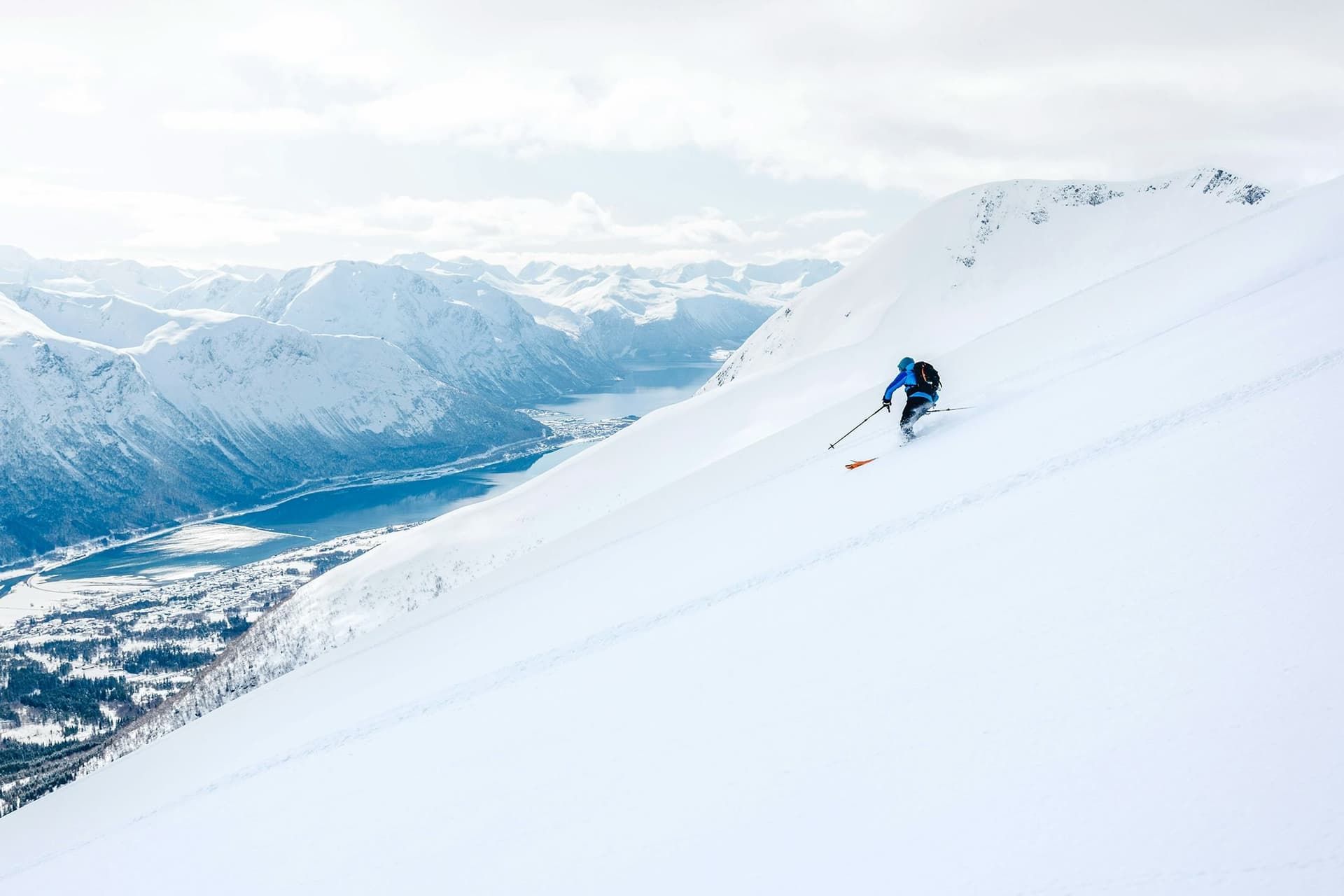 A young man skiing down a snow-covered slope in Norway, with a breathtaking view of the mountains and a fjord below, experiencing a once-in-a-lifetime adventure during his school exchange programme in Norway.