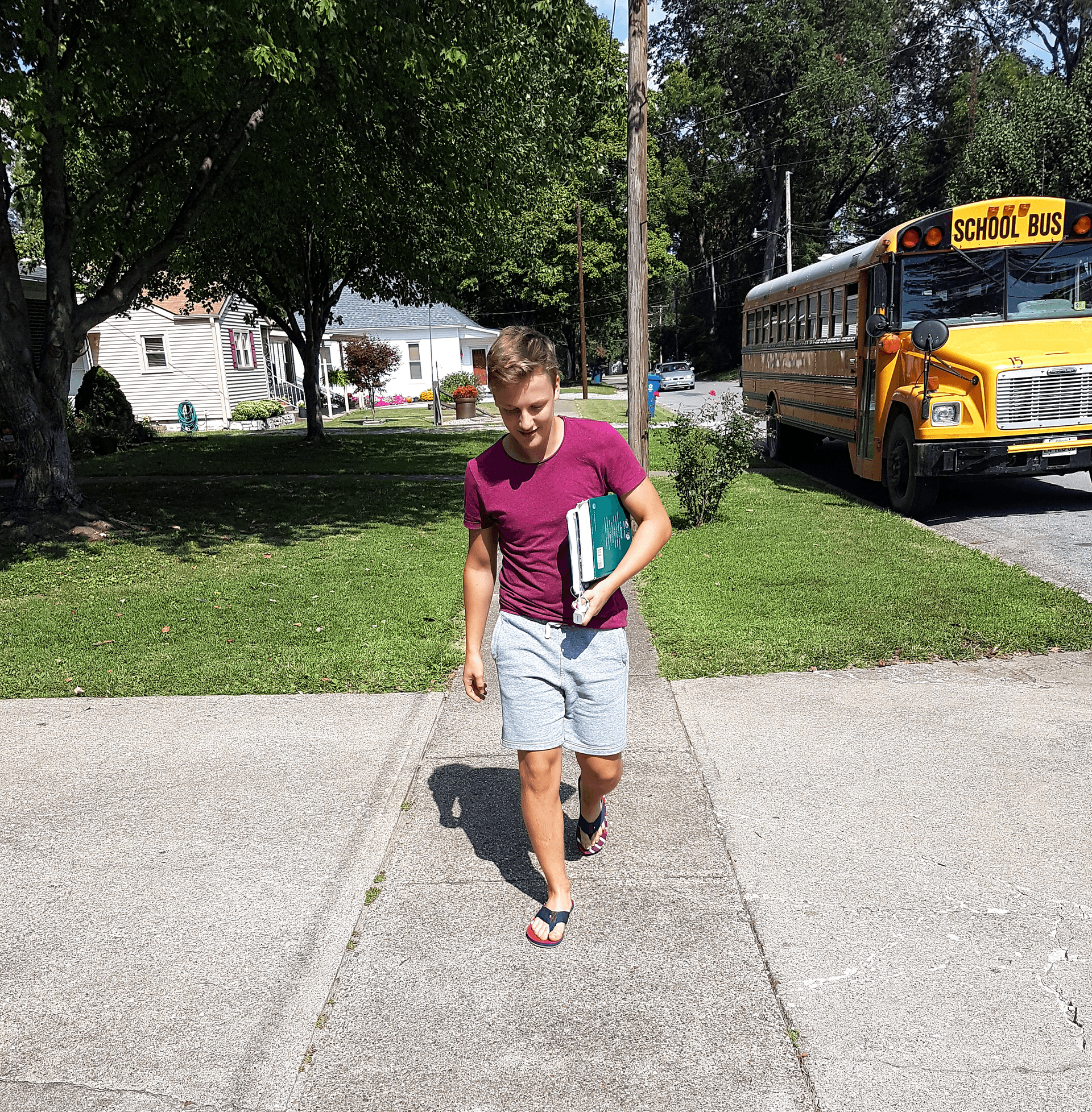 Studente cammina verso casa con libri dopo la scuola, con uno school bus sullo sfondo, scena tipica del sistema scolastico americano.