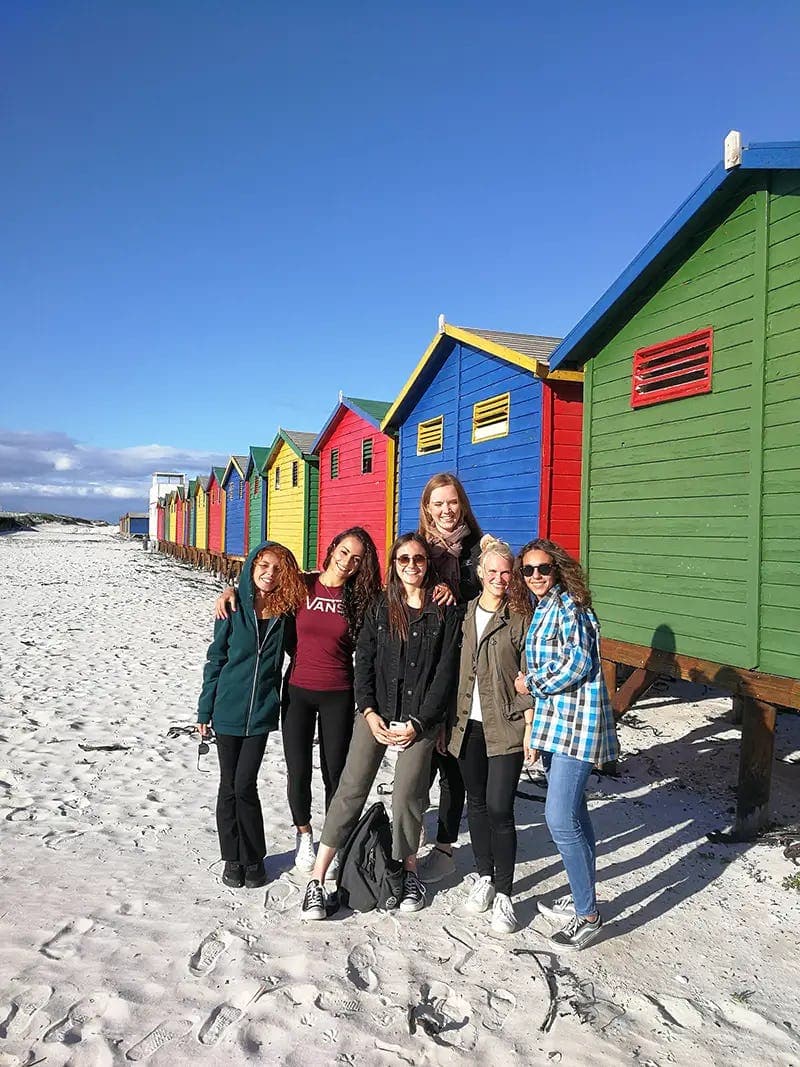 A group of students smiling on the beaches of South Africa, with colourful huts in the background, during their school exchange programme.