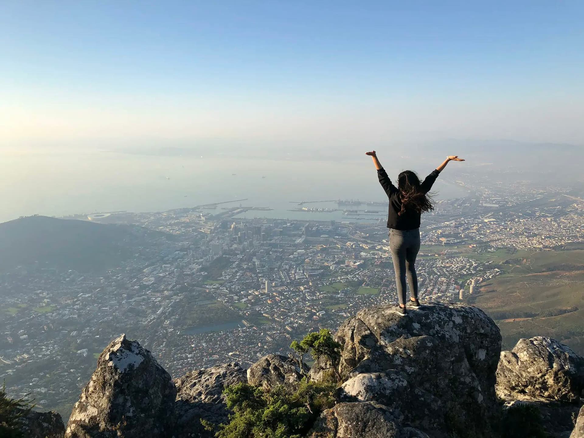 A young woman standing atop a mountain in South Africa, arms raised in triumph, enjoying a spectacular view of Cape Town during her school exchange programme in South Africa.