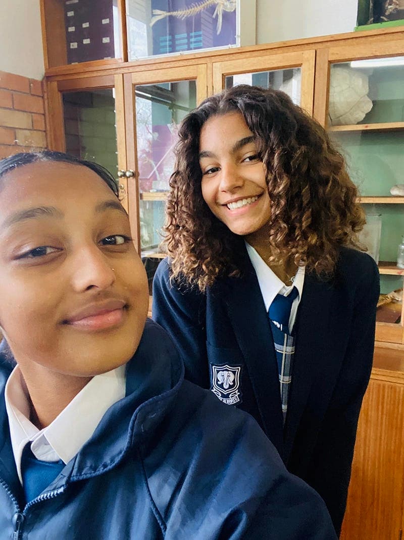 Two smiling students in school uniforms during their study year abroad in South Africa, with a local school in the background.